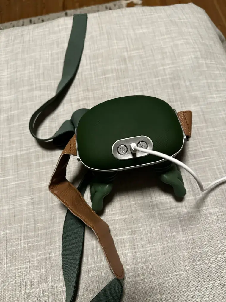 The Nodly massager resting on a linen cushion, its green and brown strap unrolled, charging cable plugged in